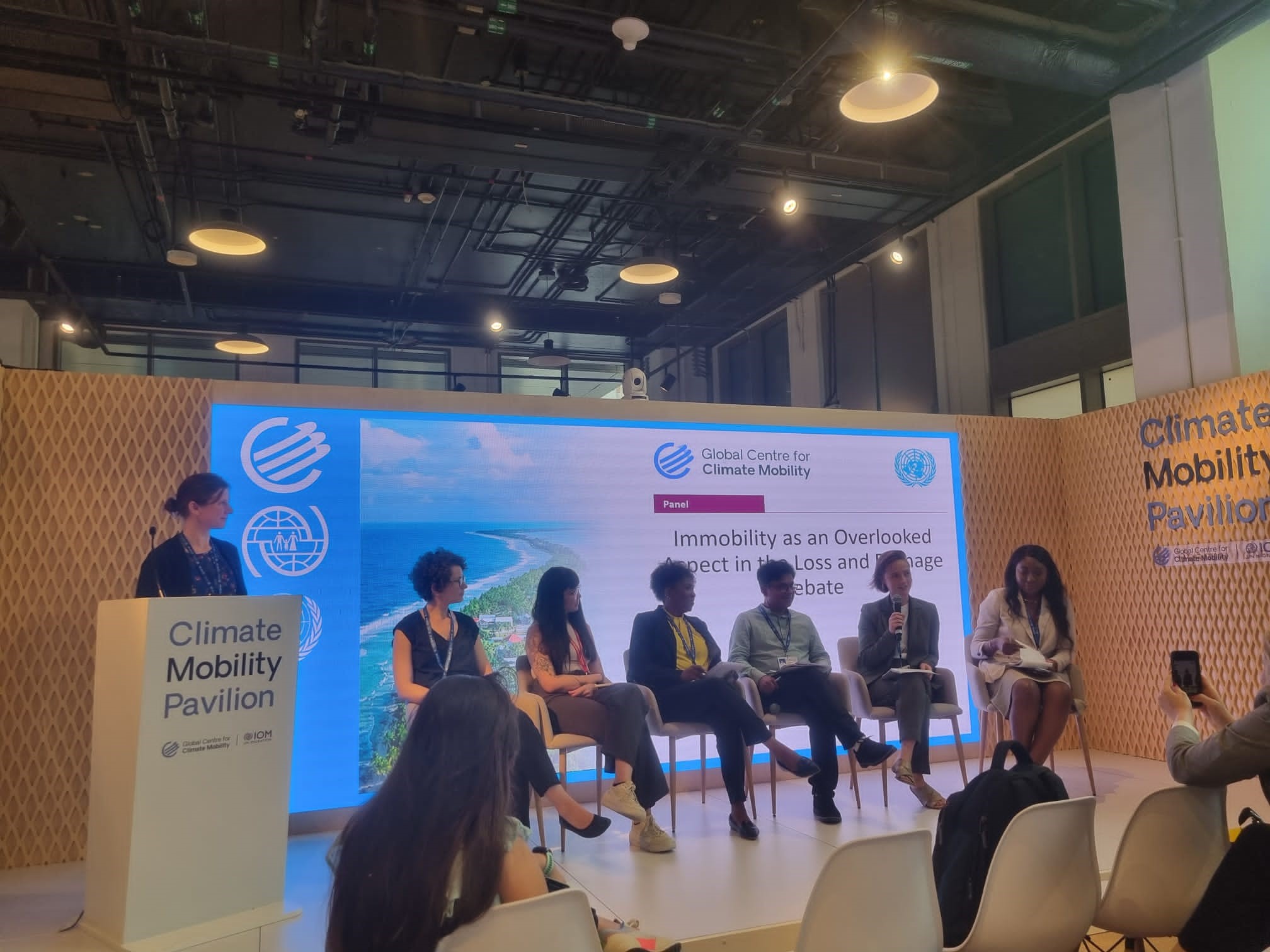 Photo: Cop28 Side Event, Panel of the 6 panelists sitting on stage and the moderator standing in front of the speakers podium, POV from the audience towards the panel talking about "Immobility as an overlooked aspect in the loss and damage debate".