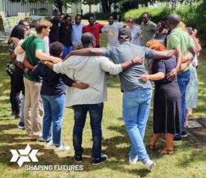 Gruppenfoto: Die Teilnehmer der Shaping Futures Academy stehen Arm in Arm in einem Kreis, während sie sich an einem sonnigen Tag im Freien aufhalten.