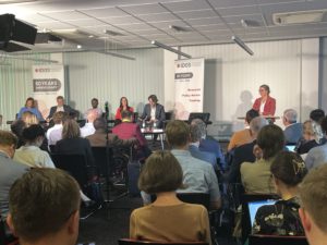 Photo: Podiumsteilnehmer*innen, darunter Julius Mbatia (African Group of Negotiators), Carola Mejía (Latindad), Jonathan Beynon (CGD) und Dr. Svea Koch (IDOS), Blickwinkel aus dem Publikum heraus zur Bühne gerichtet.