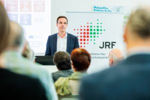 Photo: Wulf Reiners speaks to the audience in front of the logo of the Johannes Rau Research Association