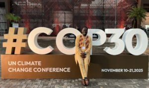 Große Installation mit weißem Schriftzug „#COP30“ auf braunem Sockel. Darunter steht „UN Climate Change Conference“ und rechts „November 10–21, 2025“. Eine Person sitzt auf dem Sockel. Hintergrund: moderne Architektur mit geometrischem Muster und Pflanzen.