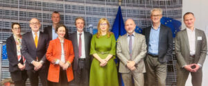 A group of people standing in an office setting in front of a wall with a grid pattern, with EU flags in the background.