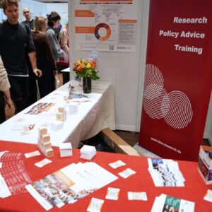 IDOS‑Messestand mit Informationsmaterialien auf Tischen, einem großen Poster mit grafischer Darstellung und einem Roll‑up mit der Aufschrift ‚Research – Policy Advice – Training‘.