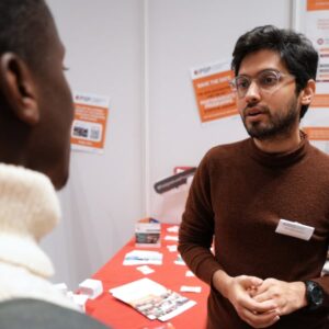 Zwei Personen stehen am IDOS‑Stand im Gespräch miteinander; im Hintergrund hängen Informationsposter und liegen Broschüren aus.