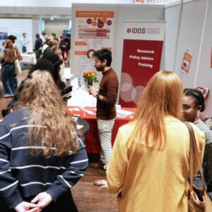 Mehrere Messebesucher*innen stehen vor dem IDOS‑Stand, an dem Informationsmaterialien ausgelegt und Poster ausgestellt sind.