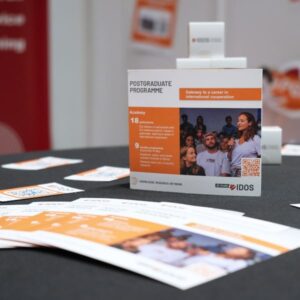 Table at the IDOS booth displaying flyers and a Postgraduate Programme information card, with additional materials visible in the background.