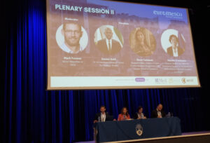 Foto: Großer Konferenzsaal mit einer Bühne. Im Hintergrund ist eine Präsentationsfolie mit dem Titel „PLENARY SESSION II“ zu sehen, darunter Porträts eines Moderators und mehrerer Diskussionsteilnehmender.