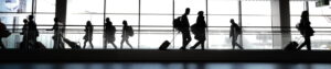 People with rolling suitcases walk through a large airport terminal, silhouetted against floor-to-ceiling windows. The scene conveys travel, mobility, and international movement.