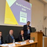 Podiumsveranstaltung beim World Trade Forum 2026. Mehrere Teilnehmende sitzen auf dem Podium, während eine Person am Mikrofon spricht. Im Hintergrund ist die Veranstaltungsgrafik zu sehen.