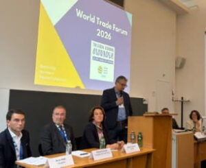 Podiumsveranstaltung beim World Trade Forum 2026. Mehrere Teilnehmende sitzen auf dem Podium, während eine Person am Mikrofon spricht. Im Hintergrund ist die Veranstaltungsgrafik zu sehen.