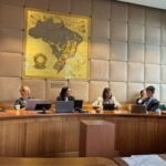 Diskussionsrunde beim WTO Public Forum in einem Konferenzraum. Mehrere Teilnehmende sitzen an einem runden Tisch vor Laptops und Unterlagen. Im Hintergrund ist eine große historische Weltkarte an der Wand zu sehen.