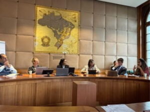 Diskussionsrunde beim WTO Public Forum in einem Konferenzraum. Mehrere Teilnehmende sitzen an einem runden Tisch vor Laptops und Unterlagen. Im Hintergrund ist eine große historische Weltkarte an der Wand zu sehen.